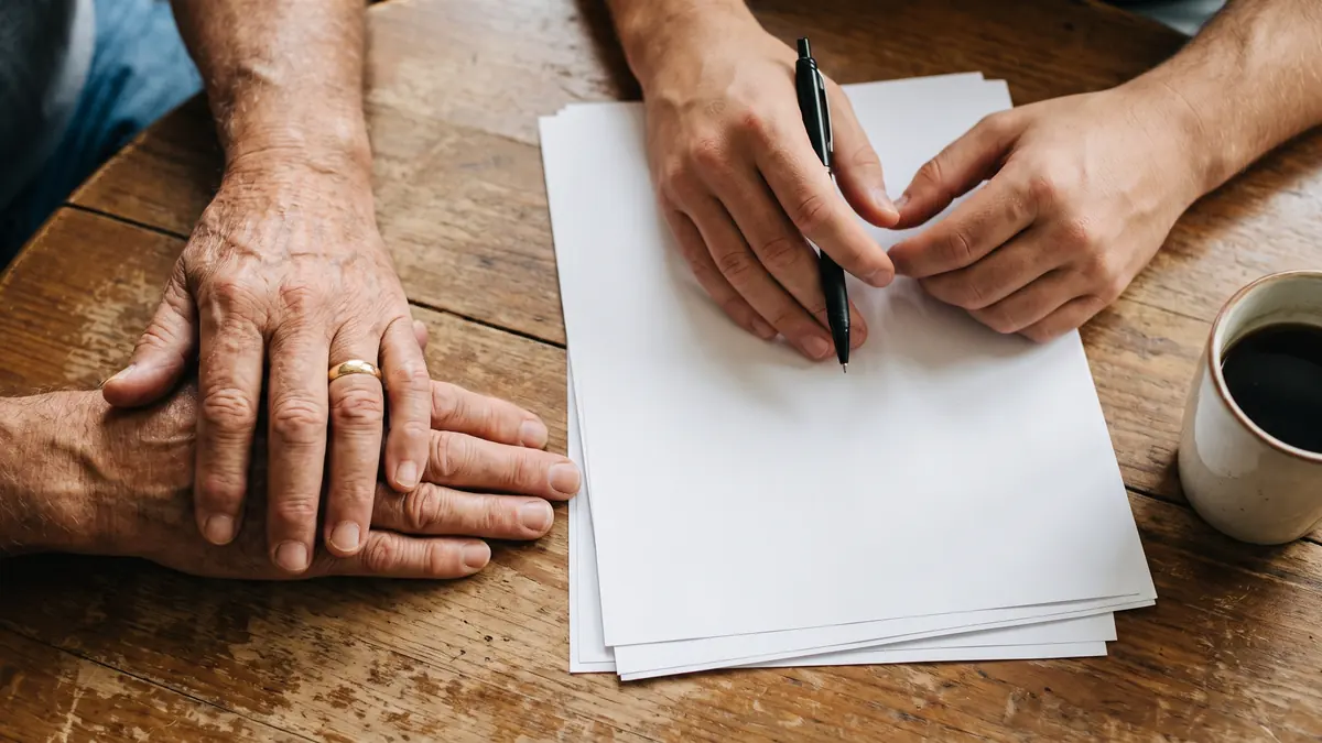 An older person’s hands and a younger adult’s hands resting on a wooden table beside papers and a pen.